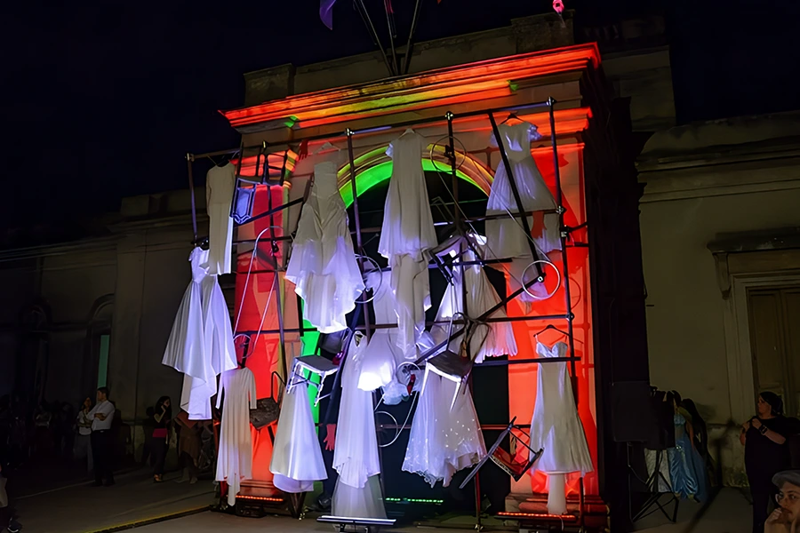 Installazione artistica con abiti bianchi sospesi su struttura metallica durante un evento serale.