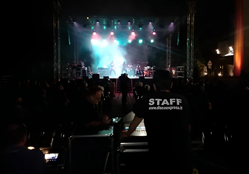 Tecnici del team Disco Express al lavoro durante un concerto, con palco illuminato e pubblico in sala.