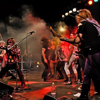 Concerto rock con band e pubblico sul palco tra luci colorate ed effetti scenici