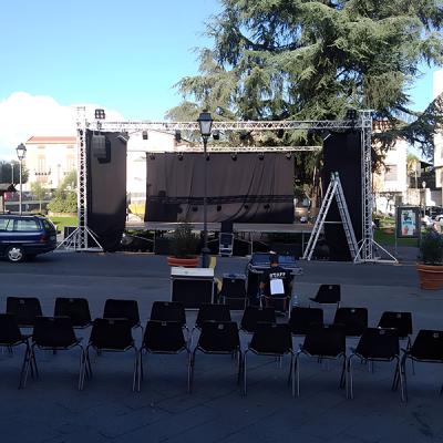 Palco allestito all’aperto in una piazza cittadina con sedie disposte per il pubblico e impianto audio in preparazione