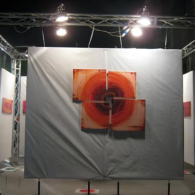 Installazione artistica con quattro tele quadrate astratte in tonalità rosse disposte a croce su un pannello grigio.