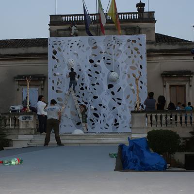 Tecnici montano un fondale scenografico traforato su un palco esterno davanti a un edificio storico.