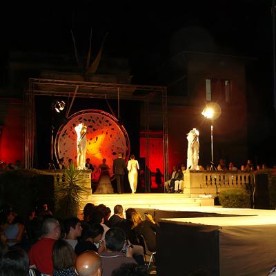 Sfilata di moda serale con passerella illuminata e pubblico seduto, scenografia rossa e struttura metallica circolare sullo sfondo.