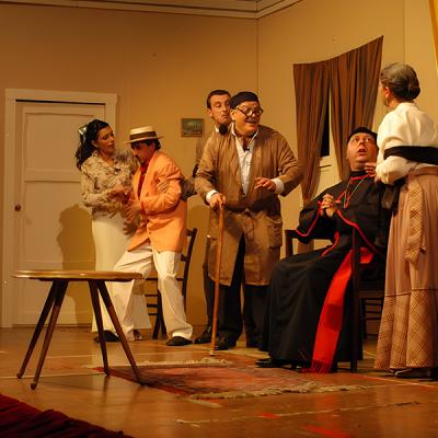 Scena teatrale comica con attori in costume d’epoca su palco di teatro, ambientazione domestica.