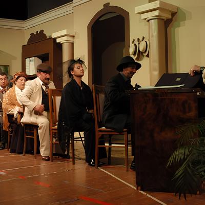 Scena teatrale d’interni con attori in abiti eleganti in fila davanti a una scrivania, commedia d’epoca.