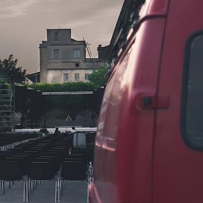 Allestimento di palco teatrale all’aperto con sedie vuote e furgone rosso in primo piano.