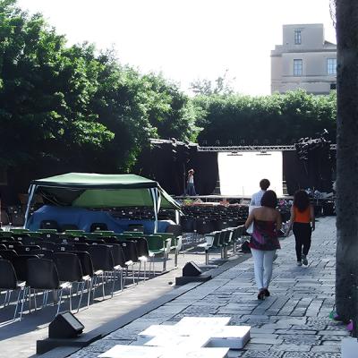 Allestimento teatrale all’aperto con palco in preparazione e persone che si avvicinano.