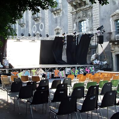 Palco teatrale allestito in piazza storica con sedie colorate e decorazioni artistiche.