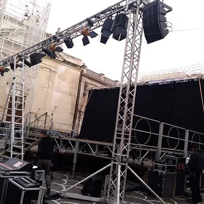 Allestimento tecnico di un palco per spettacolo teatrale all’aperto in piazza, con struttura in traliccio, impianto luci e casse audio in fase di montaggio.