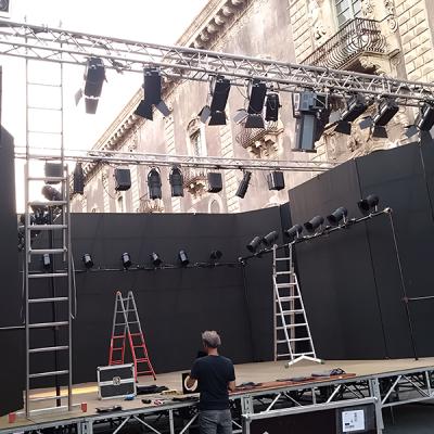 Tecnico allestisce un palco scenico all'aperto con fondale nero e impianto luci professionale in una piazza storica.