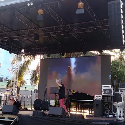 Palco allestito con LED wall e strumenti musicali per evento live all'aperto, tecnico al lavoro davanti a un pianoforte.